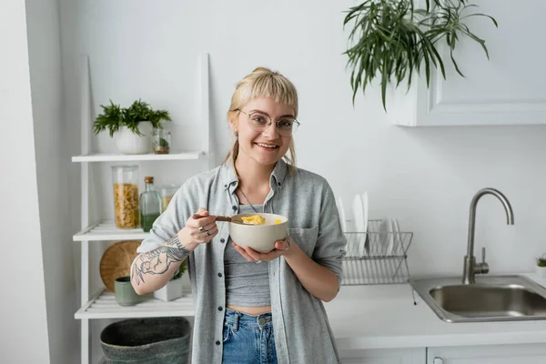 Mujer joven tatuada con flequillo y anteojos sonriendo mientras sostiene el tazón con hojuelas de maíz y cuchara mientras desayuna al lado del estante con plantas y fregadero de cocina por la mañana en un apartamento moderno - foto de stock