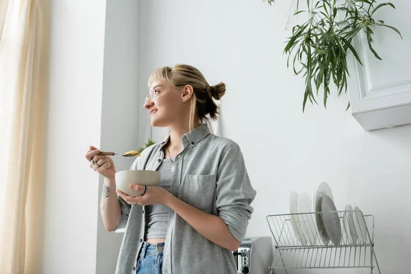 Mujer joven tatuada con flequillo y anteojos sonriendo mientras sostiene un tazón con copos de maíz y cuchara mientras desayuna y mira hacia otro lado cerca de plantas verdes y platos limpios en cocina blanca - foto de stock