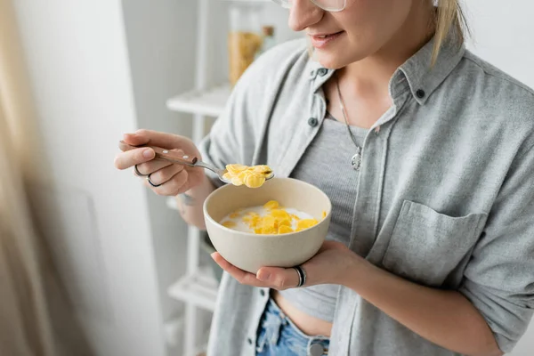Cropped view of tattooed young woman smiling while holding bowl with cornflakes and spoon while having breakfast next to blurred white rack in modern kitchen. copy space, apartment, morning energy — Stock Photo