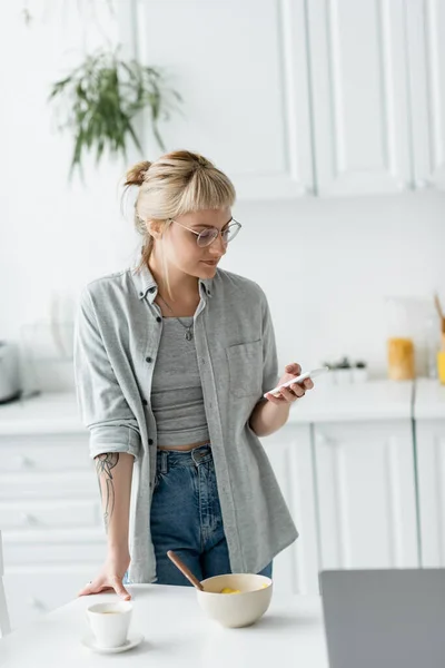 Young woman with short hair, tattoo and bangs using smartphone while standing in eyeglasses near bowl with cornflakes, cup of coffee and laptop on table in modern apartment, freelancer — Stock Photo