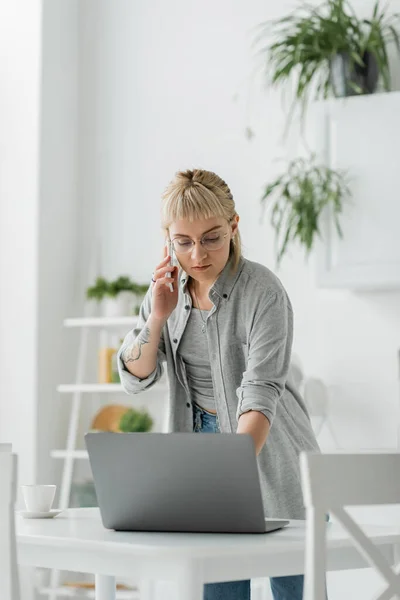 Young woman with short hair, bangs and tattoo on hand talking on smartphone while standing in eyeglasses near cup of coffee and laptop on table in modern apartment, freelancer — Stock Photo