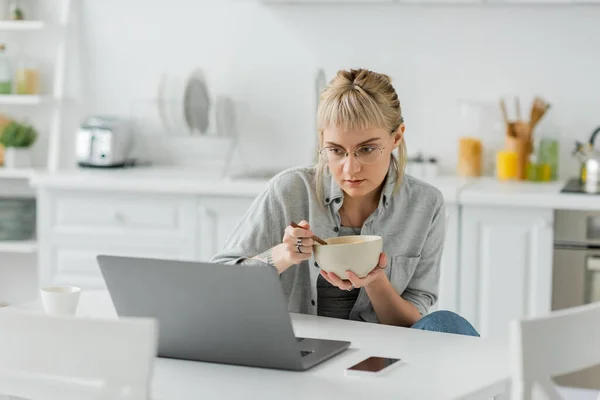 Молодая женщина с челкой и татуировкой на руке едят кукурузные хлопья на завтрак при использовании ноутбука рядом со смартфоном с чистым экраном и чашкой кофе на столе в современной кухне, фрилансер — стоковое фото