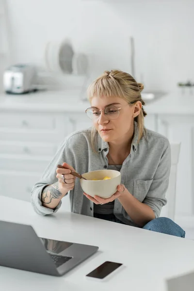 Молода жінка з чубчиком і татуюванням на руці їсть кукурудзяні пластівці на сніданок, дивлячись на ноутбук біля смартфона з порожнім екраном на столі в сучасній кухні, фрілансер, працює з дому — стокове фото