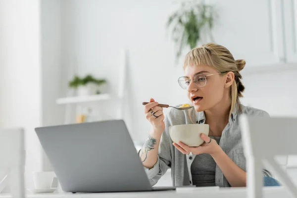 Mujer joven con flequillo y tatuaje en la mano comer copos de maíz para el desayuno mientras se utiliza el ordenador portátil cerca de teléfono inteligente y una taza de café en la mesa en la cocina moderna, freelancer - foto de stock