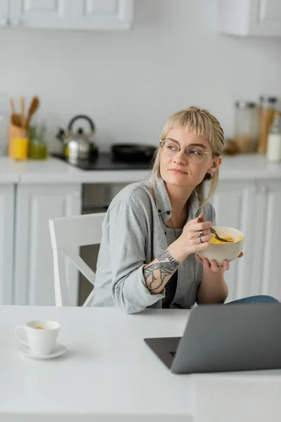 Mujer joven soñadora con flequillo, pelo corto y tatuaje en la mano comer copos de maíz para el desayuno, mientras que el uso de la computadora portátil cerca de la taza de café con platillo en la mesa en la cocina moderna, freelancer, trabajo desde casa - foto de stock
