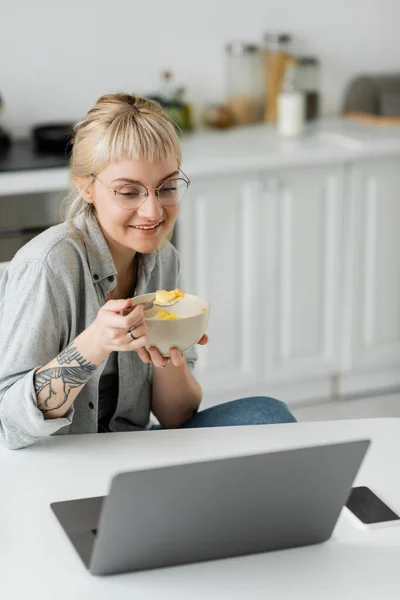 Счастливая молодая женщина с короткими волосами, челка и татуировка на руке кукурузные хлопья на завтрак во время использования ноутбука рядом со смартфоном с чистым экраном на столе и улыбаясь на современной кухне, фрилансер — стоковое фото
