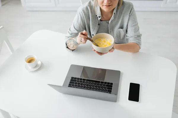 Vista ritagliata di giovane donna con tatuaggio a portata di mano mangiare cornflakes per la prima colazione durante l'utilizzo di laptop vicino smartphone con schermo bianco e una tazza di caffè sul tavolo in cucina moderna, libero professionista — Foto stock