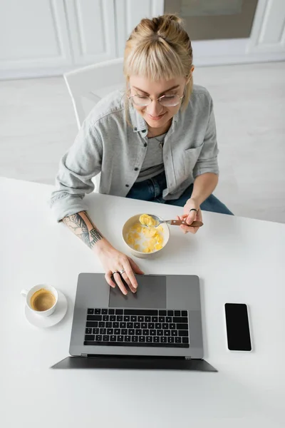 Vista superior de la joven feliz con tatuaje en la mano comer copos de maíz para el desayuno mientras se utiliza el ordenador portátil cerca de teléfono inteligente con pantalla en blanco y taza de café en la mesa en la cocina moderna, freelancer - foto de stock
