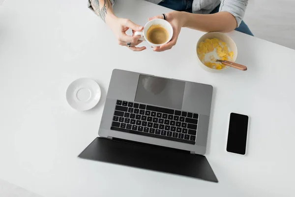 Top view of woman with tattoo on hand holding cup of coffee near bowl with cornflakes during breakfast while using laptop near smartphone with blank screen in modern kitchen, freelancer, cropped — Stock Photo