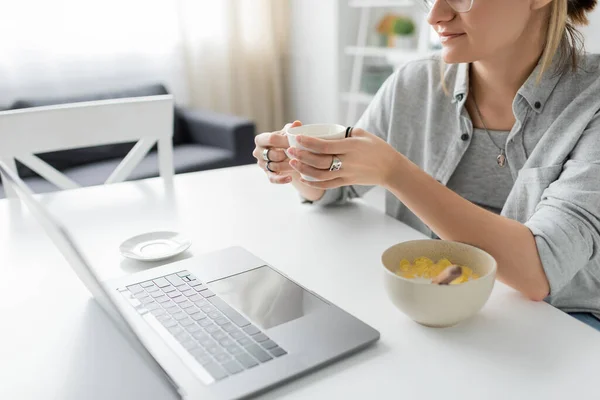Plan recadré de jeune femme tenant une tasse de café près d'un bol avec des flocons de maïs pendant le petit déjeuner tout en utilisant un ordinateur portable près de la soucoupe blanche sur la table dans la cuisine moderne, pigiste, travail de la maison — Photo de stock