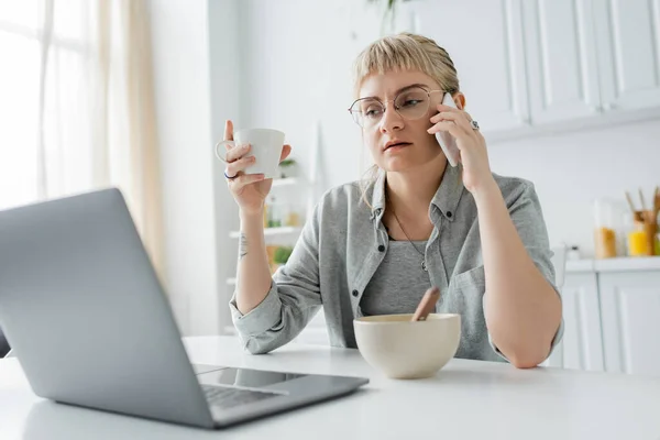 Young woman with tattoo on hand and bangs talking on smartphone while using laptop, holding cup of coffee near bowl wth cornflakes on table in modern kitchen, freelancer, work from home — Stock Photo