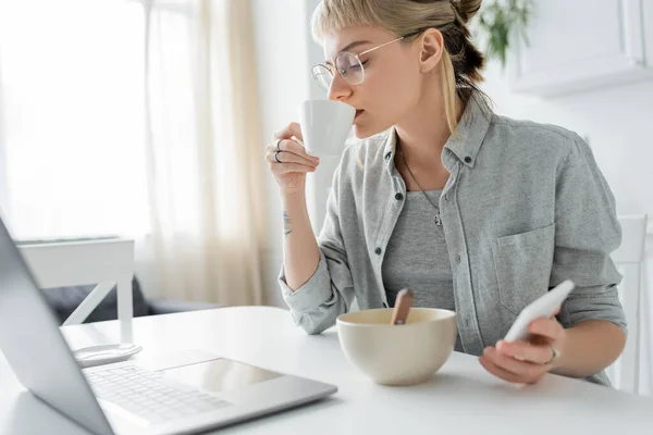 Mujer joven con tatuaje en la mano y flequillo que sostiene el teléfono inteligente y el uso de la computadora portátil, beber café mientras sostiene la taza cerca y bowl wth copos de maíz en la mesa en la cocina moderna, freelancer - foto de stock