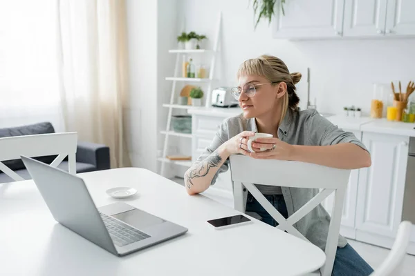 Mujer joven con tatuaje en la mano y flequillo sosteniendo taza de café y mirando a la computadora portátil cerca de teléfono inteligente y platillo en la mesa blanca alrededor de sillas en la cocina moderna, freelancer, estilo de vida remoto - foto de stock