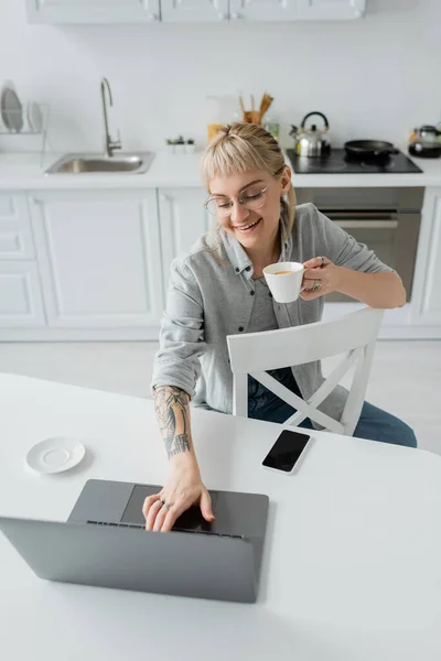 Vista aérea de la joven feliz con tatuaje en la mano y flequillo sosteniendo la taza de café y mirando a la computadora portátil cerca de teléfono inteligente y platillo en la mesa blanca alrededor de sillas en la cocina moderna, estilo de vida remoto - foto de stock