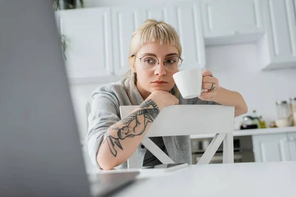 Enfocada mujer joven con tatuaje en la mano y flequillo sosteniendo taza de café y mirando borrosa portátil cerca de teléfono inteligente en la mesa blanca en la cocina moderna, freelancer, estilo de vida remoto - foto de stock