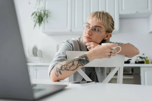 Dreamy young woman with tattoo on hand and bangs holding cup of coffee and looking away near blurred laptop and smartphone on white table in modern kitchen, freelancer, remote lifestyle — Stock Photo