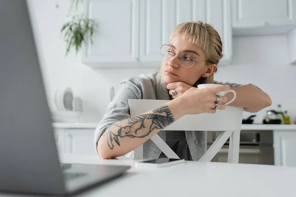 Verträumte junge Frau mit Tätowierung auf der Hand und Pony, Kaffeebecher haltend und in der Nähe von verschwommenem Laptop und Smartphone auf weißem Tisch in moderner Küche, Freiberuflerin, abgelegener Lebensstil — Stockfoto