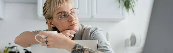 Dreamy young woman with tattoo on hand and bangs holding cup of coffee and looking away near blurred laptop in modern kitchen, freelancer, remote lifestyle, eyeglasses, banner — Stock Photo
