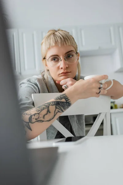 Giovane donna annoiata in occhiali con tatuaggio a portata di mano e frangia in possesso di tazza di caffè e guardando il computer portatile vicino smartphone su tavolo bianco in cucina moderna, freelance, stile di vita remoto — Foto stock