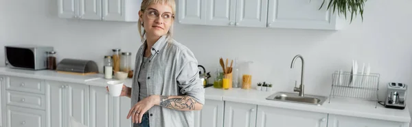 Mujer joven en gafas con tatuaje en la mano y flequillo sosteniendo la taza de café y mirando hacia otro lado mientras está de pie en la cocina moderna con diferentes electrodomésticos y gabinetes blancos, pancarta, hora de la mañana - foto de stock