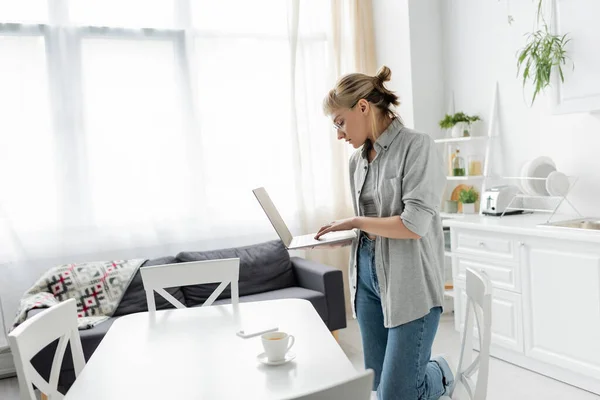 Young woman in eyeglasses with short hair and bangs holding laptop near cup of coffee and smartphone on white table around chairs in white and modern kitchen next to sofa, freelancer, remote lifestyle — Stock Photo