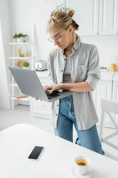 Mujer joven en gafas con pelo corto y flequillo sosteniendo portátil cerca de taza de café y teléfono inteligente con pantalla en blanco en la mesa blanca en la cocina blanca y moderna, estilo de vida remoto, freelancer - foto de stock