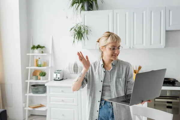 Happy young woman in eyeglasses, short hair and bangs holding laptop and waving hand during video call in white and modern kitchen, blurred background, remote lifestyle, freelancer — Stock Photo