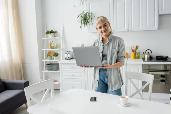 Mujer joven feliz en gafas con el pelo corto y flequillo que sostiene el ordenador portátil y mirando a la cámara cerca de la taza de café y teléfono inteligente con pantalla en blanco en la mesa blanca y cocina moderna, freelance - foto de stock