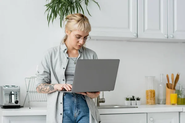 Tattooed young woman in eyeglasses, short hair and bangs holding and using laptop while working from home in white and modern kitchen, blurred background, remote lifestyle, freelancer — Stock Photo