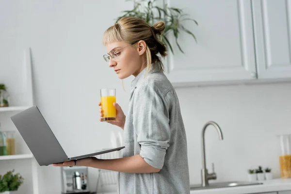 Young woman in eyeglasses, short hair and bangs holding glass of fresh orange juice and using laptop while working from home in white and modern kitchen, blurred background, freelancer — Stock Photo