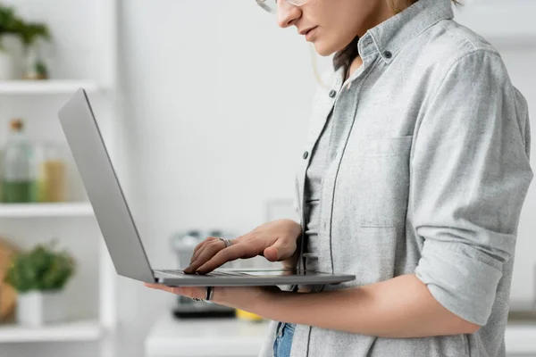 Vista cortada de focada jovem mulher em cinza camisa segurando e usando laptop na cozinha branca e moderna, fundo borrado, estilo de vida remoto, freelancer, trabalho em casa, autônomo — Fotografia de Stock
