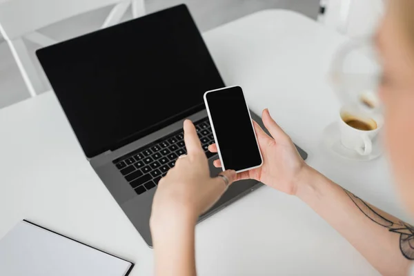 Vue du haut d'une femme tatouée tenant un smartphone avec écran vierge près d'un ordinateur portable, d'un ordinateur portable et d'une tasse de café avec soucoupe sur une table blanche tout en travaillant à la maison, pigiste, espace de travail moderne — Photo de stock