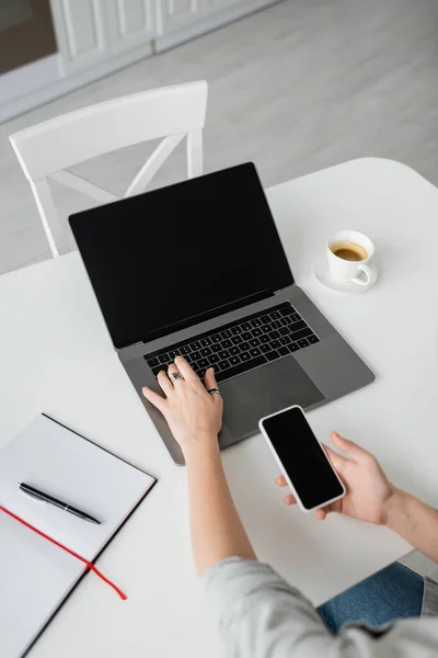 Вид сверху на женщину, держащую смартфон с чистым экраном и использующую ноутбук рядом с ноутбуком с ручкой, и чашку кофе с блюдцем на белом столе во время работы из дома, фрилансер, современное рабочее пространство — стоковое фото
