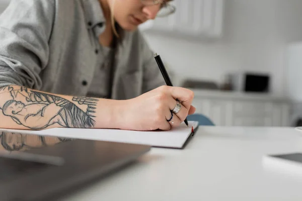 Vista recortada de la mujer joven con tatuaje en la escritura de la mano en el cuaderno, tomando notas, teniendo inspiración mientras sostiene la pluma cerca del ordenador portátil en la mesa blanca, primer plano borroso, trabajo desde casa - foto de stock