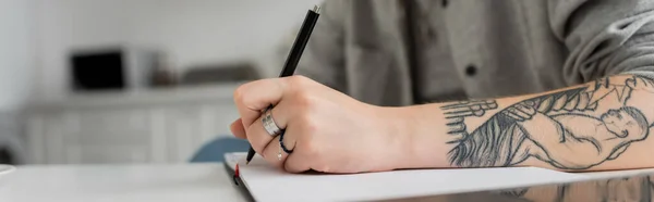 Cropped shot of young woman with tattoo on hand writing in notebook, taking notes, having inspiration while holding pen near laptop on white table, blurred foreground, work from home, banner — Stock Photo