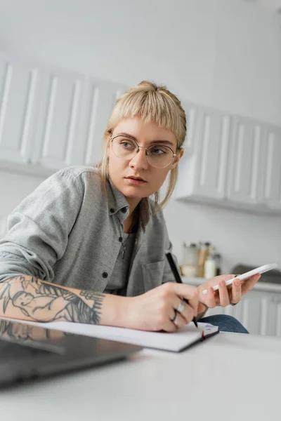 Young woman with tattoo on hand writing in notebook, taking notes, holding smartphone and pen near laptop on white table, blurred foreground, work from home — Stock Photo