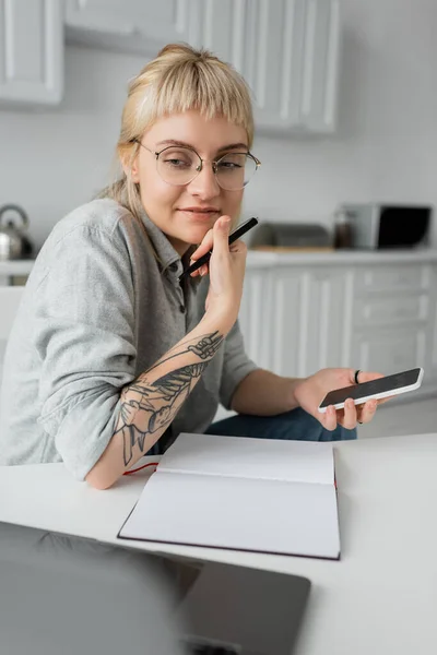 Щаслива молода жінка з татуюванням на руці і чубчиком тримає смартфон з порожнім екраном і ручкою біля ноутбука і ноутбука на білому столі, розмитим переднім планом, працює з дому — стокове фото
