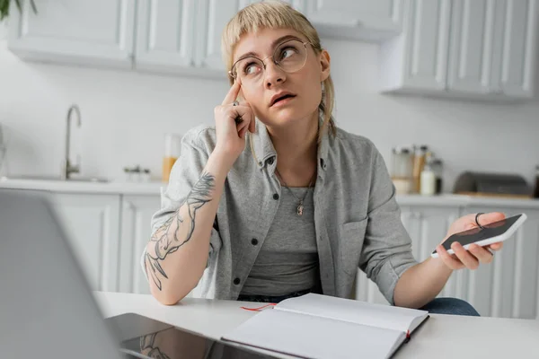Pensive young woman with tattoo on hand and bangs holding smartphone with blank screen and pen near notebook and laptop on white table, blurred foreground, work from home — Stock Photo