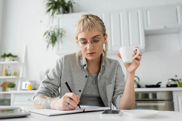 Mujer joven con tatuaje en la mano y flequillo sosteniendo taza de café cerca de portátil, teléfono inteligente y portátil en la mesa blanca, primer plano borroso, trabajo desde casa, tomando notas - foto de stock