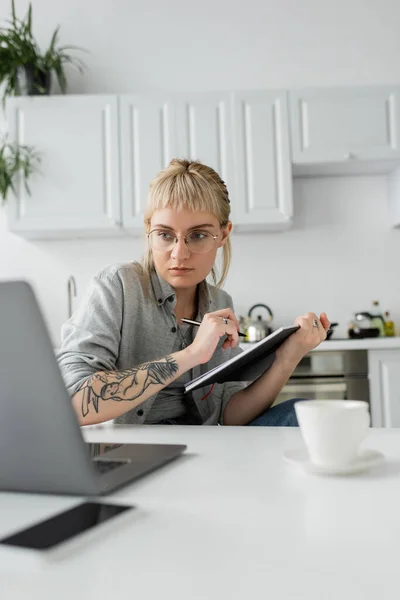 Молода жінка з татуюванням на руці і чубчиком тримає блокнот, роблячи нотатки біля смартфона і ноутбука на білому столі, розмитий передній план, працює з дому — стокове фото