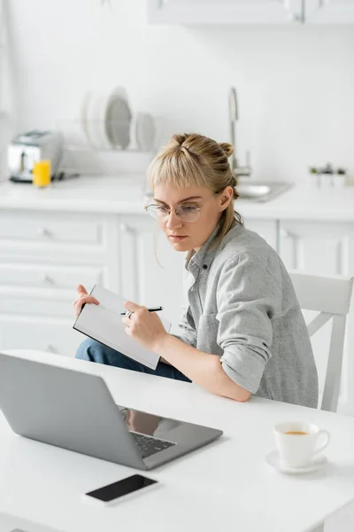 Young woman with tattoo on hand and bangs holding notebook, taking notes near smartphone and laptop on white table, blurred background , work from home — Stock Photo