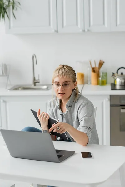 Молодая женщина с татуировкой на руке и челкой, держащая ноутбук, рядом смартфон с чистым экраном и глядя на ноутбук на белом столе, размытый передний план, работа из дома — стоковое фото