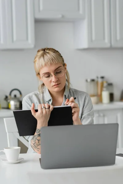 Mujer joven en gafas con tatuaje en la mano y flequillo sosteniendo cuaderno, tomando notas, sentado cerca de la computadora portátil y taza de café en la mesa blanca, fondo borroso, trabajo desde casa - foto de stock