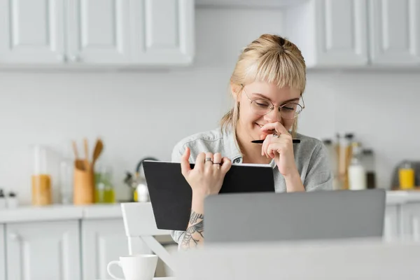 Happy young woman in eyeglasses with tattoo on hand and bangs laughing, covering mouth, holding notebook, taking notes, sitting near laptop and cup of coffee on table, blurred background, — Stock Photo