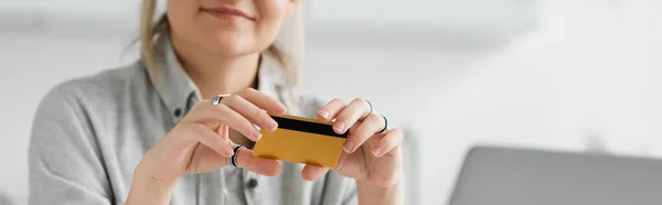 Cropped view of happy young woman smiling and holding credit card, sitting near blurred laptop in modern apartment, workspace, digital lifestyle,online transactions, technology, banner — Stock Photo