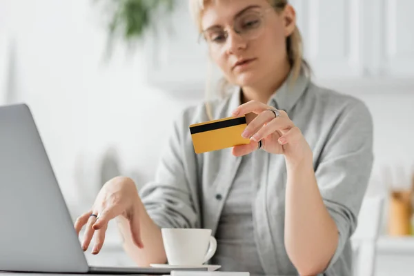 Mujer joven en gafas con tatuaje en la mano con tarjeta de crédito, sentado cerca de la computadora portátil y una taza de café en la mesa blanca, fondo borroso, trabajo desde casa, transacciones en línea, tecnología - foto de stock