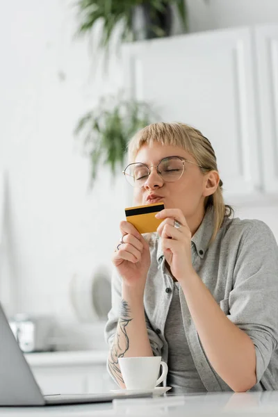 Jeune femme en lunettes avec tatouage sur la main embrassant carte de crédit, assis près d'un ordinateur portable et tasse de café sur une table blanche, fond flou, travail de la maison, transactions en ligne, la technologie — Photo de stock