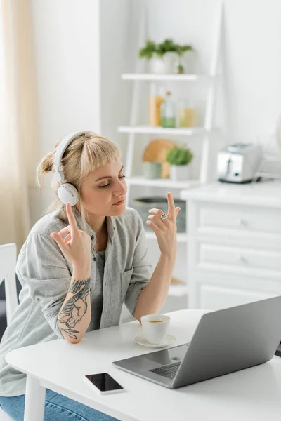 Young woman with bangs and tattoo on hand sitting in wireless headphones and gesturing near laptop, cup of coffee and blurred smartphone with blank screen on table, freelance, work from home — Stock Photo