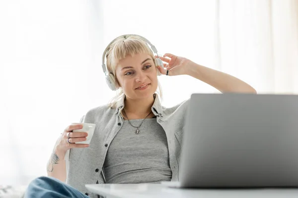Smiling young woman with bangs and tattoo on hand sitting in wireless headphones and holding cup of coffee while looking at laptop, on blurred table, freelance, work from home — Stock Photo