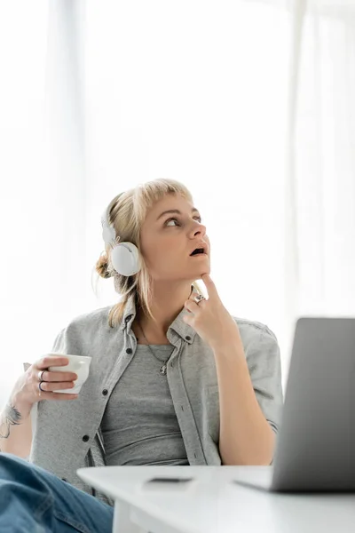 Chère jeune femme aux cheveux blonds, frange et tatouage à portée de main assise dans un casque sans fil et tenant une tasse de café près d'un ordinateur portable et un smartphone flou sur la table. freelance. travail à domicile — Photo de stock